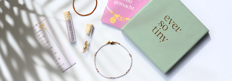 Flatlay eines DIY-Schmucksets von ever so tiny mit Perlenröhrchen, Draht, Maßband und grün-goldener Box – stilvoll arrangiert im Sonnenlicht.