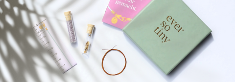 Flatlay eines DIY-Schmucksets von ever so tiny mit Perlenröhrchen, Draht, Maßband und grün-goldener Box – stilvoll arrangiert im Sonnenlicht.