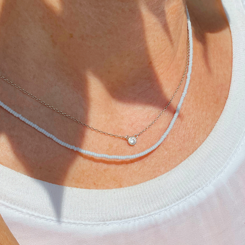 Nahaufnahme eines Dekolletés, auf dem zwei zarte Halsketten getragen werden. Die untere Kette besteht aus kleinen, pastellblauen japanischen Glasperlen, während die obere Kette eine feine, silberne Gliederkette mit einem einzelnen, funkelnden Steinanhänger ist. Beide Ketten sind minimalistisch und ergänzen sich stilvoll, ideal für einen modernen, dezenten Look.