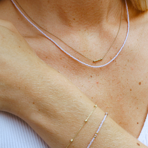 Nahaufnahme eines Dekolletés mit zwei zarten Halsketten. Die erste Kette besteht aus kleinen, lavendelfarbenen Glasperlen, die zweite ist eine feine goldene Gliederkette mit kleinen goldenen Perlen als Akzent. Dazu wird ein passendes Armband aus einer Kombination von lavendelfarbenen Glasperlen und einer goldenen Kette getragen. Der Look ist elegant und dezent, ideal für einen stilvollen und persönlichen Stil.