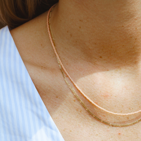 Nahaufnahme eines Dekolletés mit zwei zarten Halsketten. Die obere Kette besteht aus kleinen, peachfarbenen Glasperlen, die untere ist eine feine goldene Gliederkette. Die Kombination aus den peachfarbenen Perlen und der Goldkette verleiht dem Look eine schlichte Eleganz und passt perfekt zu sommerlicher Kleidung. Ideal für einen minimalistischen, stilvollen Auftritt.