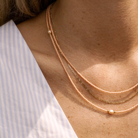 Eine Frau trägt drei filigrane Halsketten in zarten Pfirsich- und Goldtönen. Die Ketten bestehen aus feinen Glasperlen, goldenen Akzenten und einer einzelnen Süßwasserperle als Highlight. Der elegante Lagenlook der Ketten harmoniert perfekt mit dem gestreiften Oberteil in Blau und Weiß und verleiht dem Outfit eine stilvolle und sommerliche Note. Ideal für DIY-Schmuckliebhaber, die einen minimalistischen und hochwertigen Look bevorzugen.