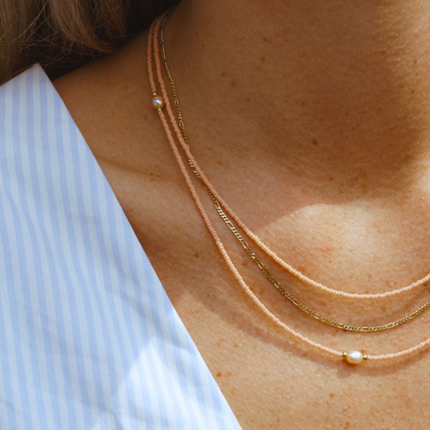 Nahaufnahme eines Dekolletés mit drei übereinander getragenen Halsketten. Zwei der Ketten bestehen aus peachfarbenen Glasperlen, eine davon ist mit einer einzelnen Süßwasserperle als Highlight verziert. Die dritte Kette ist eine feine goldene Gliederkette. Diese Kombination aus Perlen und Gold verleiht dem Look eine elegante und feminine Note, ideal für einen stilvollen, sommerlichen Auftritt.