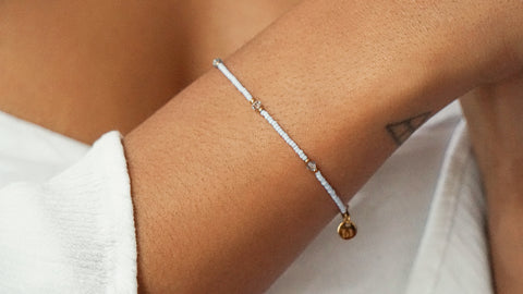 Zartes DIY-Armband aus hellblauen Glasperlen mit goldfarbenen Details und Swarovski-Elementen, zum selber machen mit nachhaltigen Materialien von ever so tiny. Getragen am Handgelenk, kombiniert mit einem weißen Oberteil.