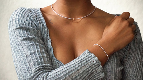 Frau trägt ein DIY-Schmuckset bestehend aus einer zarten Halskette und einem passenden Armband in Lavendelfarben mit goldfarbenen Details und Süßwasserperlen, zum selber machen mit nachhaltigen Materialien von ever so tiny. Kombiniert mit einem gerippten, hellgrauen Oberteil.