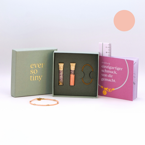 DIY-Schmuckset von ever so tiny für ein Armband in zartem Apricot mit goldfarbenen Details und Swarovski-Kristallen. Das Set wird in einer eleganten, jadegrünen Box mit hochwertiger Goldprägung präsentiert und enthält alles, was für die Schmuckherstellung benötigt wird: Glasperlen, Verschluss, Werkzeug und eine Anleitung für einfaches Selbermachen. Im Vordergrund ist ein fertiges Beispielarmband zu sehen.