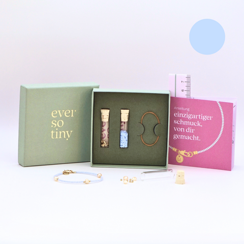 DIY-Schmuckset von ever so tiny für ein Armband in Hellblau mit goldfarbenen Details und Süßwasserperlen. Präsentiert in einer edlen, jadegrünen Box mit Goldprägung, enthält das Set Glasperlen, Verschluss und eine Schritt-für-Schritt-Anleitung. Im Vordergrund sind ein fertiges Beispielarmband und die enthaltenen Materialien zu sehen.