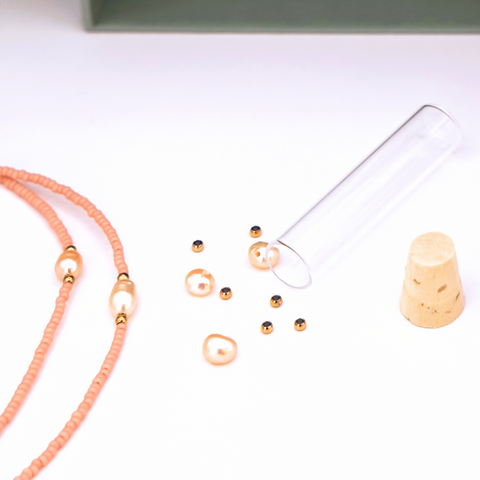 Close-Up auf DIY-Schmuckset von ever so tiny für eine Halskette in zartem Peach mit goldfarbenen Details und Süßwasserperlen. Präsentiert in einer hochwertigen, jadegrünen Box mit edler Goldprägung. Das Set enthält Glasperlen, Verschluss und eine Schritt-für-Schritt-Anleitung zur einfachen Herstellung. Im Vordergrund ist ein fertiges Beispiel der Kette zu sehen.