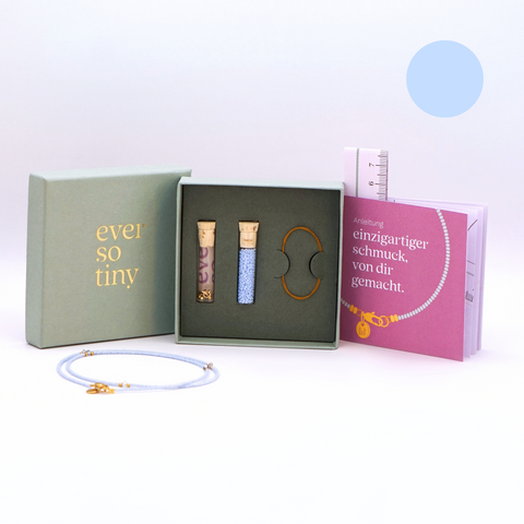 DIY-Schmuckset von ever so tiny für eine Halskette in Hellblau mit goldfarbenen Details und Swarovski-Kristallen. Das Set wird in einer nachhaltigen, jadegrünen Box mit edler Goldprägung präsentiert und enthält Glasperlen, Verschluss,  sowie eine Schritt-für-Schritt-Anleitung für die einfache Schmuckherstellung. Im Vordergrund ist ein fertiges Beispiel der Kette zu sehen.
