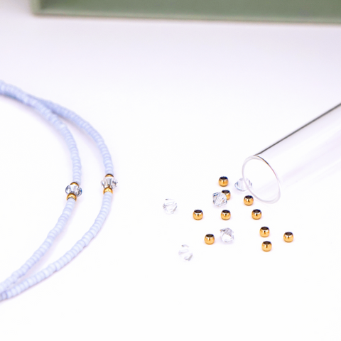 Close-Up auf DIY-Schmuckset von ever so tiny für eine Halskette in Hellblau mit goldfarbenen Details und Swarovski-Kristallen. Das Set wird in einer nachhaltigen, jadegrünen Box mit edler Goldprägung präsentiert und enthält Glasperlen, Verschluss,  sowie eine Schritt-für-Schritt-Anleitung für die einfache Schmuckherstellung. Im Vordergrund ist ein fertiges Beispiel der Kette zu sehen.