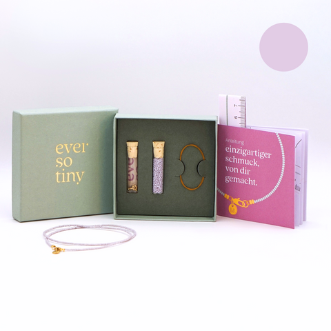 DIY-Schmuckset von ever so tiny mit lavendelfarbenen Glasperlen, goldfarbenem Verschluss und Zubehör, präsentiert in einer nachhaltigen, jadefarbenen Schmuckbox mit hochwertiger Goldprägung des Logos. Enthält eine Anleitung mit Schritt-für-Schritt-Erklärungen zur einfachen Schmuckherstellung und ein fertiges Beispiel einer Kette in Lavendel.