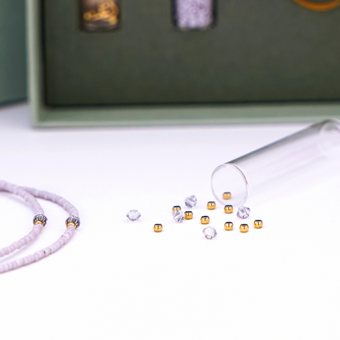 Close-Up auf DIY-Schmuckset von ever so tiny für eine Halskette in Lavendelfarben mit goldfarbenen Details und Swarovski-Kristallen. Präsentiert in einer hochwertigen, jadegrünen Box mit edler Goldprägung. Das Set enthält Glasperlen, Verschluss und eine Schritt-für-Schritt-Anleitung für die einfache Schmuckherstellung. Im Vordergrund ist ein fertiges Beispiel der Kette zu sehen.