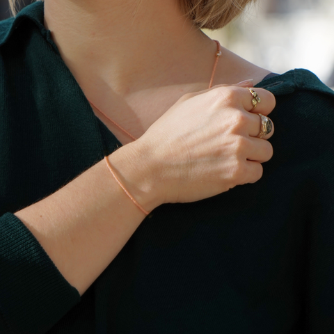 Eine Person trägt ein peach farbened filigranes Perlenarmband, welches sie selbst hergestellt hat. Sie trägt einen dunkelgrünen Pullover, hat blondes, mittellanges Haar, an ihrem Dekolletee sieht man eine dazu passende Kette mit einer Süßwasserperle eingearbeitet. Zudem trägt sie zwei hochwertige goldene Ringe.