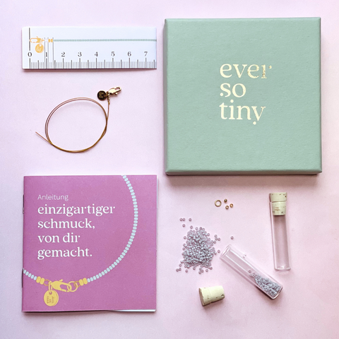 Ein DIY-Schmuckset von „ever so tiny“, bestehend aus einer mintfarbenen Schmuckbox mit goldener Prägung, goldfarbenem Schmuckdraht, einer lila Anleitung, Maßband, Glasperlen in einem Röhrchen mit Korkverschluss und kleinen Zubehörteilen.
DIY-Schmuckset von „ever so tiny“: Hochwertige Materialien wie Glasperlen, goldfarbener Schmuckdraht und nachhaltige Verpackung. Enthält Anleitung und Zubehör für einzigartigen, selbstgemachten Schmuck. Ideal für kreative Projekte und besondere Gesche