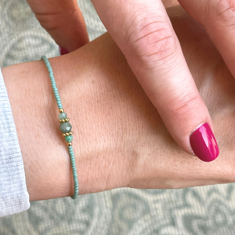 Das Bild zeigt ein filigranes Armband, welches von einer Person mit rotem Nagellack auf den Fingern getragen wird. In der Mitte des Armbands befindet sich ein facettierter, tiefgrüner Smaragd, der von zwei goldfarbenen Spacer-Perlen eingerahmt wird. Diese sind 18K goldplattiert und verleihen dem Schmuckstück einen edlen Akzent. Die restlichen Perlen sind feine, jadefarbene japanische Glasperlen mit gleichmäßiger Form und zarter Farbgebung.
