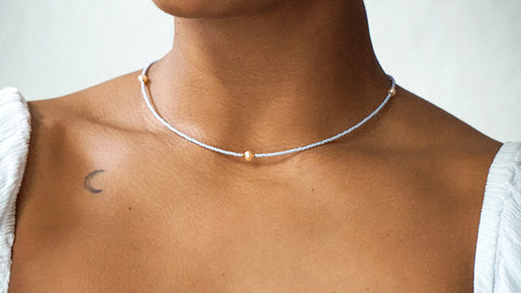 Zarte DIY-Halskette aus hellblauen Glasperlen mit goldfarbenen Details und einer Süßwasserperle als Highlight, ideal zum Selbermachen mit dem Schmuckset von ever so tiny. Getragen zu einem weißen Oberteil.