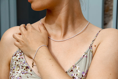 Eine Frau trägt eine zarte Halskette und ein Armband aus kleinen, pastellblauen Glasperlen mit goldfarbenen Akzenten. Die Hand ruht leicht auf der Schulter, was die schlichte Eleganz der Schmuckstücke betont. Die Schmuckstücke sind dezent und passen perfekt zu dem sommerlichen Outfit mit floralen Mustern. Ideal für einen minimalistischen und stilvollen Look.