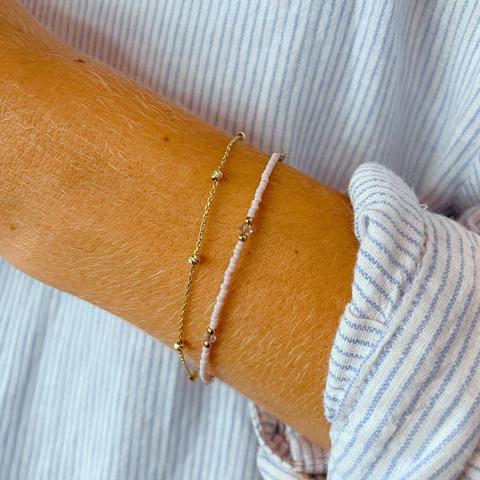 Eine Frau trägt eine schlichte Halskette und ein passendes Armband, beide aus kleinen, lavendelfarbenen Glasperlen. Das Armband ist mit einem goldfarbenen Anhänger verziert. Die dezenten Schmuckstücke betonen den eleganten, minimalistischen Stil und passen ideal zu sommerlicher Kleidung. Perfekt für einen dezenten und stilvollen Look.