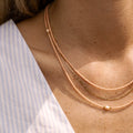 Eine Frau trägt drei filigrane Halsketten in zarten Pfirsich- und Goldtönen. Die Ketten bestehen aus feinen Glasperlen, goldenen Akzenten und einer einzelnen Süßwasserperle als Highlight. Der elegante Lagenlook der Ketten harmoniert perfekt mit dem gestreiften Oberteil in Blau und Weiß und verleiht dem Outfit eine stilvolle und sommerliche Note. Ideal für DIY-Schmuckliebhaber, die einen minimalistischen und hochwertigen Look bevorzugen.