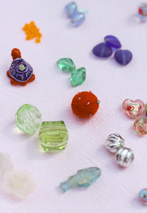 Verschiedene Perlen aus Glas, Filz und Plastik in bunten Formen und Farben auf rosa Stoff. Symbolisiert die Vielfalt an Materialien und Designs für kreative Schmuckprojekte.