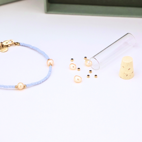 Close-Up auf DIY-Schmuckset von ever so tiny für ein Armband in Hellblau mit goldfarbenen Details und Süßwasserperlen. Präsentiert in einer edlen, jadegrünen Box mit Goldprägung, enthält das Set Glasperlen, Verschluss und eine Schritt-für-Schritt-Anleitung. Im Vordergrund sind ein fertiges Beispielarmband und die enthaltenen Materialien zu sehen.