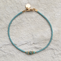 Das Bild zeigt ein filigranes Armband, das auf einer hellen, natürlichen Oberfläche liegt. In der Mitte des Armbands befindet sich ein facettierter, tiefgrüner Smaragd, der von zwei goldfarbenen Spacer-Perlen eingerahmt wird. Diese sind 18K goldplattiert und verleihen dem Schmuckstück einen edlen Akzent. Die restlichen Perlen sind feine, jadefarbene japanische Glasperlen mit gleichmäßiger Form und zarter Farbgebung. 