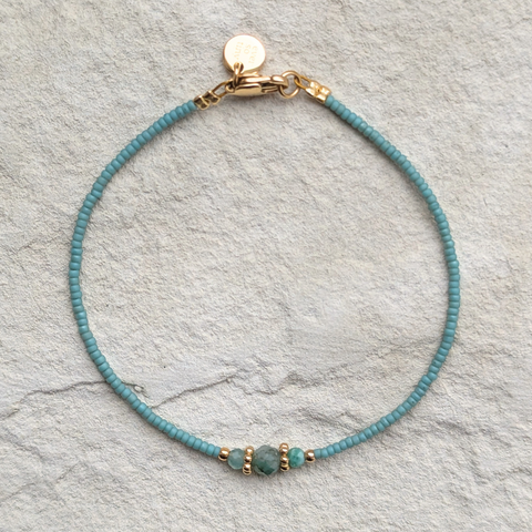 Das Bild zeigt ein filigranes Armband, das auf einer hellen, natürlichen Oberfläche liegt. In der Mitte des Armbands befindet sich ein facettierter, tiefgrüner Smaragd, der von zwei goldfarbenen Spacer-Perlen eingerahmt wird. Diese sind 18K goldplattiert und verleihen dem Schmuckstück einen edlen Akzent. Die restlichen Perlen sind feine, jadefarbene japanische Glasperlen mit gleichmäßiger Form und zarter Farbgebung. 