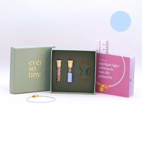 DIY-Schmuckset von ever so tiny mit hellblauen Glasperlen, goldfarbenem Verschluss und Zubehör, präsentiert in einer nachhaltigen, jadefarbenen Schmuckbox mit hochwertiger Glodprägung des Logos. Enthält eine Anleitung mit Schritt-für-Schritt-Erklärungen zur einfachen Schmuckherstellung und ein fertiges Beispielarmband in blau. 