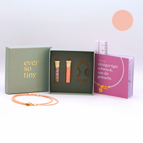 DIY-Schmuckset von ever so tiny für eine Halskette in zartem Peach mit goldfarbenen Details und Süßwasserperlen. Präsentiert in einer hochwertigen, jadegrünen Box mit edler Goldprägung. Das Set enthält Glasperlen, Verschluss und eine Schritt-für-Schritt-Anleitung zur einfachen Herstellung. Im Vordergrund ist ein fertiges Beispiel der Kette zu sehen.