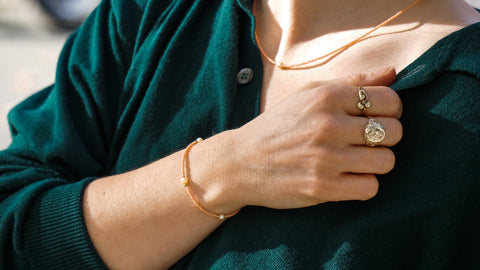 Filigranes Armband in Peach mit recycelten Süßwasserperlen, zart am Handgelenk getragen, kombiniert mit goldenen Ringen und einem grünen Pullover.