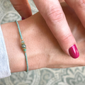 Das Bild zeigt ein filigranes Armband, welches von einer Person mit rotem Nagellack auf den Fingern getragen wird. In der Mitte des Armbands befindet sich ein facettierter, tiefgrüner Smaragd, der von zwei goldfarbenen Spacer-Perlen eingerahmt wird. Diese sind 18K goldplattiert und verleihen dem Schmuckstück einen edlen Akzent. Die restlichen Perlen sind feine, jadefarbene japanische Glasperlen mit gleichmäßiger Form und zarter Farbgebung. 